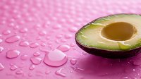 Fresh cut avocado half with pit on wet pink surface with water drops, minimal food composition for healthy eating and cosmetic concepts.