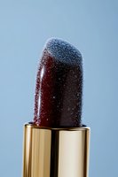 Dark burgundy lipstick with sparkling glitter texture in golden tube against soft blue background for luxury cosmetics and makeup concepts.