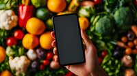 Hand holding smartphone with blank black screen over colorful fresh vegetables and fruits background for food delivery app mockup or grocery shopping.