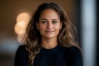Young Hispanic woman with long wavy brown hair wearing black sweater smiling warmly at camera against blurred background with soft bokeh lights.