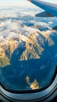 Aerial view of snow-capped alpine mountains with scattered clouds and blue wing of aircraft in frame, captured from airplane window during flight over mountainous terrain.