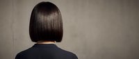 Woman with straight black bob hairstyle viewed from behind wearing dark top against neutral gray background for hair care and salon concepts.