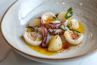 Grilled octopus tentacles served with baby potatoes and quail eggs in olive oil dressing, garnished with fresh herbs and paprika on ceramic plate. Fine dining presentation.