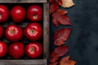 Fresh red apples in rustic wooden box with vibrant autumn leaves on dark background, seasonal harvest composition for food and fall decoration.