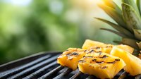 Grilled pineapple slices with caramelized grill marks on black barbecue grate against blurred green background, fresh summer tropical fruit cooking outdoors.