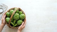 Female hands holding wicker basket filled with fresh green artichokes on white textured surface, overhead view for food styling and culinary content.