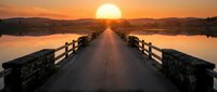 Wooden bridge stretching toward golden sunset over calm water, symmetrical perspective with warm orange sky and distant mountains, serene landscape for travel and nature themes.
