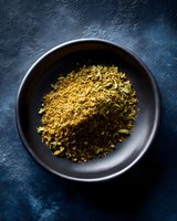 Ground dried herbs and spices mix in black ceramic bowl on dark blue textured background, overhead view for culinary and food photography.