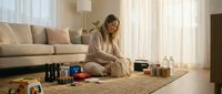 Young woman in cozy pajamas sitting on living room floor organizing skincare and beauty products for morning routine in bright natural light.
