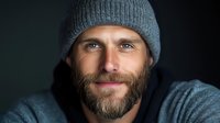 Handsome bearded man in gray knit beanie hat smiling warmly at camera against dark background. Portrait of confident casual male model for lifestyle content.