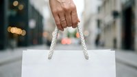 Person holding white shopping bag with rope handles on urban street with blurred city background, suitable for retail, commerce, and consumer lifestyle content.