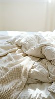 Unmade bed with crumpled beige and white bedding, rumpled duvet and textured blanket on mattress, cozy bedroom interior with neutral tones and soft natural lighting.