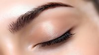 Close-up macro shot of closed eye with long eyelashes and perfectly shaped eyebrow showing professional makeup artistry and beauty treatment results.