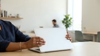 Closeup of male professional with dark skin tone working on laptop at desk in modern office environment with colleague in background.