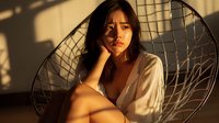 Young Asian woman in white shirt sitting in papasan chair during golden hour sunlight, creating dramatic shadows and warm atmosphere. Natural beauty portrait.