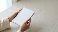 Person with medium skin tone holding blank white card or brochure mockup on neutral wooden table surface for marketing or branding purposes.