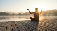 Young man practicing yoga meditation on wooden dock at sunrise over misty lake with arms raised in peaceful morning atmosphere for wellness and mindfulness.