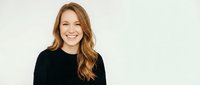 Young Caucasian woman with long wavy brown hair smiling warmly in black sweater against white background. Professional headshot for business content.