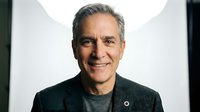 Middle-aged man with gray hair and light skin tone wearing dark gray shirt, smiling at camera against white background for corporate headshot or professional portrait use.