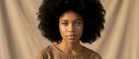 Young African American woman with natural curly afro hair wearing beige sweater against neutral backdrop with soft lighting for portrait photography.
