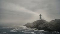 Dramatic lighthouse stands on rocky cliff overlooking stormy ocean waves under moody overcast sky creating atmospheric coastal seascape scene.