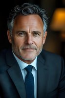 Portrait of mature Caucasian businessman with gray hair and beard wearing dark suit and blue tie, confident smile against blurred background. Professional headshot.