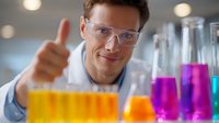 Young Caucasian male scientist in lab coat and safety glasses showing thumbs up gesture while working with colorful liquid samples in glass beakers for chemical research.
