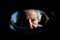 Elderly person's face reflected in rearview mirror of car, dramatic lighting emphasizes wrinkles and weathered features against dark background, creating emotional portrait.
