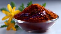 Ground red paprika powder in glass bowl with yellow flowers and fresh green herbs, macro food photography on light background.