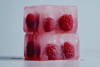 Two stacked ice cubes with frozen raspberries inside, melting on a white surface, vibrant red berries visible through clear ice.