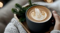 Cozy winter coffee moment with latte art rosetta pattern in black mug, knitted sweater, and pine branches against soft bokeh lights background.