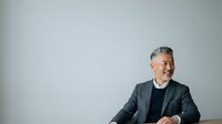 Middle-aged man with gray hair and facial hair, appearing to be of South Asian or Middle Eastern descent, wearing a dark blazer over a black shirt, seated against a neutral gray background, suitable for corporate, business, and professional contexts.