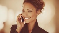 Young mixed-race woman with updo hairstyle smiling while talking on smartphone, warm bokeh background creates professional business atmosphere.