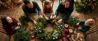 Family of five making a Christmas wreath together at a wooden table, decorating with dried oranges, cinnamon, and greenery in a festive setting.