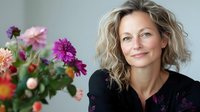 Middle-aged woman with wavy blonde hair and fair complexion, wearing black clothing, posed next to vibrant pink and purple gerbera daisies. Suitable for lifestyle, beauty, wellness, and floral design projects.