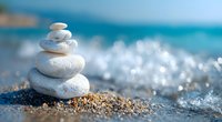 Balanced white stones stacked on sandy beach with gentle waves and turquoise ocean in soft focus background creating zen and peaceful atmosphere.