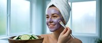 Young woman applying clay face mask with brush in bright bathroom, wearing white towel on head, skincare routine and spa treatment at home.