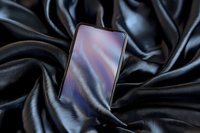 Modern smartphone with blank screen display lying on luxurious black silk fabric with elegant folds and reflective surface creating dramatic lighting effect.