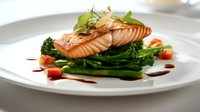 Grilled salmon fillet served on bed of steamed broccolini and asparagus, garnished with fresh herbs and balsamic reduction on white plate. Restaurant quality plating.