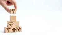Hand placing wooden block with caduceus symbol on pyramid of healthcare icons representing medical insurance and protection services.
