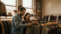 Young couple shopping together browsing clothing racks in trendy boutique store retail fashion lifestyle casual wear denim jacket smiling happy relationship.