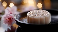 Traditional mooncake with intricate floral pattern on dark plate beside pink cherry blossoms with warm bokeh lights creating festive atmosphere.