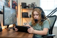 Young blonde woman gaming streamer with headphones and microphone broadcasting live gameplay session from home setup with monitor and gaming accessories.