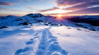 Stunning winter sunrise over snow-covered mountain peaks with footprint trail leading through pristine white landscape creating dramatic golden light rays.