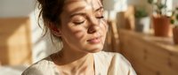 Middle-aged woman with East Asian ethnicity, wearing casual beige linen shirt, smiling with eyes closed in peaceful moment. Warm natural lighting in modern home interior with wooden furniture and potted plants. Suitable for wellness, lifestyle, and mindfulness content.