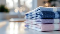 Neatly folded stack of fresh cotton towels in soft pastel blue and pink colors on white surface with blurred bokeh background, creating cozy bathroom atmosphere.