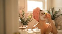 Young woman with towel on head applying skincare product in bright bathroom with plants enjoying morning beauty routine for wellness and self-care lifestyle.