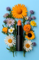 Orange lipstick surrounded by fresh summer flowers including daisies, sunflowers, and purple wildflowers on bright blue background for beauty and cosmetic marketing.