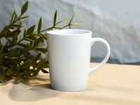 White ceramic coffee mug on wooden surface with green plant leaves in soft natural lighting, creating minimalist composition for lifestyle and beverage photography.