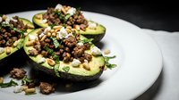 Halved avocados topped with seasoned ground meat, pine nuts, and fresh herbs served on white plate for healthy lunch or dinner meal preparation.
